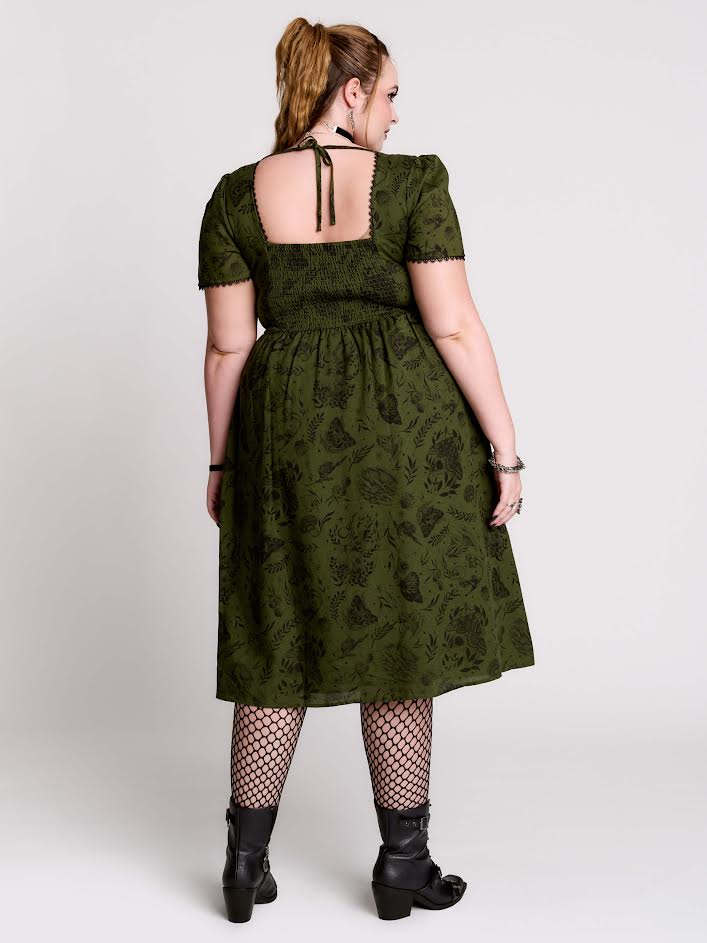 A woman shown from the back wearing an olive green short-sleeve midi dress with a dark floral jacquard pattern, a smocked bodice, and tie-back straps, paired with black fishnet tights and black ankle boots against a light gray studio background.
