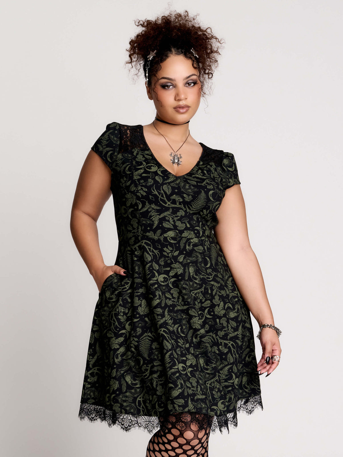 short black dress with a green leafy lace pattern, featuring cap sleeves, a V-neckline, side pockets, and a scalloped lace hem.