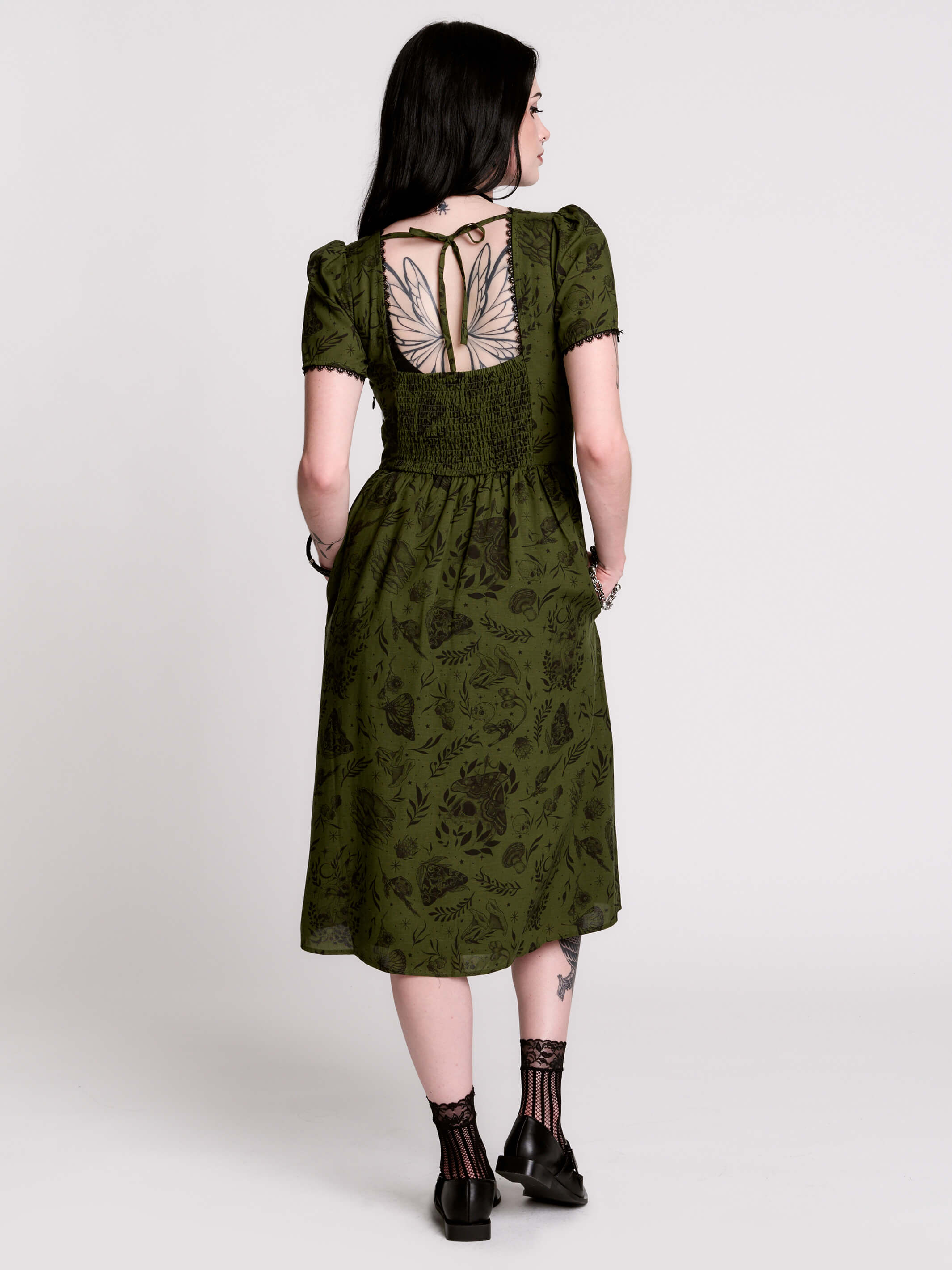 A woman viewed from the back wearing an olive green midi dress with a smocked bodice, short puff sleeves, and a tie-back neckline. The dress has a dark botanical pattern.
