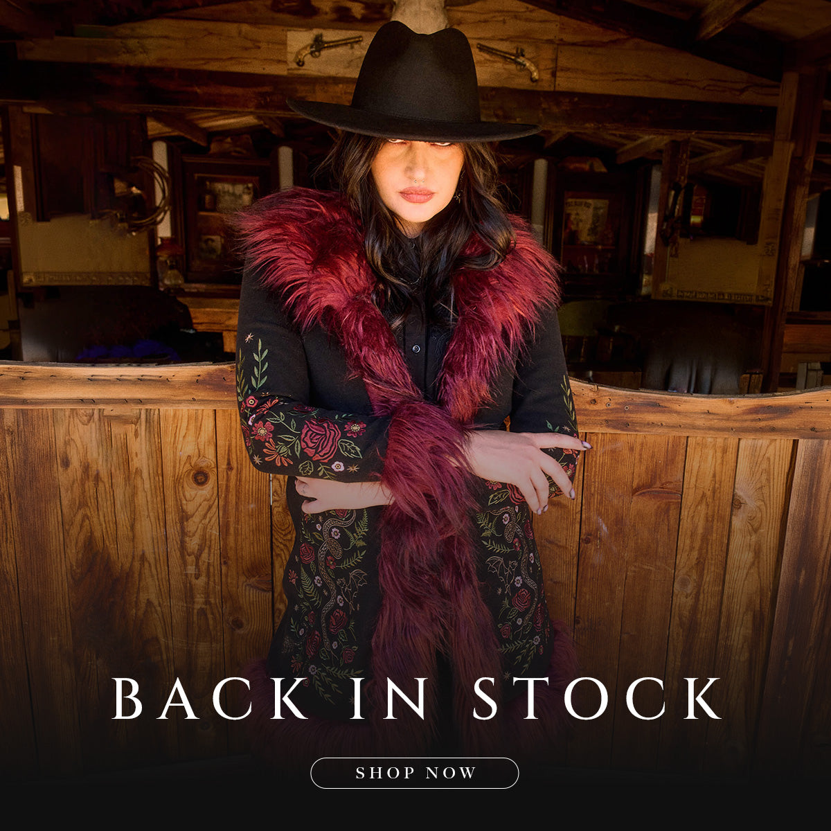 A woman wearing a black wide-brim hat, a black embroidered coat with a deep burgundy faux-fur collar, and a matching burgundy scarf, standing in a rustic wooden stable. Text overlays at the bottom read 'BACK IN STOCK' and 'SHOP NOW'.
