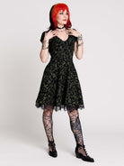 short black dress with a green leafy lace pattern, featuring cap sleeves, a V-neckline, side pockets, and a scalloped lace hem.