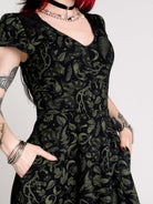 short black dress with a green leafy lace pattern, featuring cap sleeves, a V-neckline, side pockets, and a scalloped lace hem.