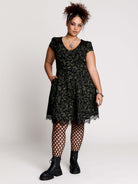 short black dress with a green leafy lace pattern, featuring cap sleeves, a V-neckline, side pockets, and a scalloped lace hem.