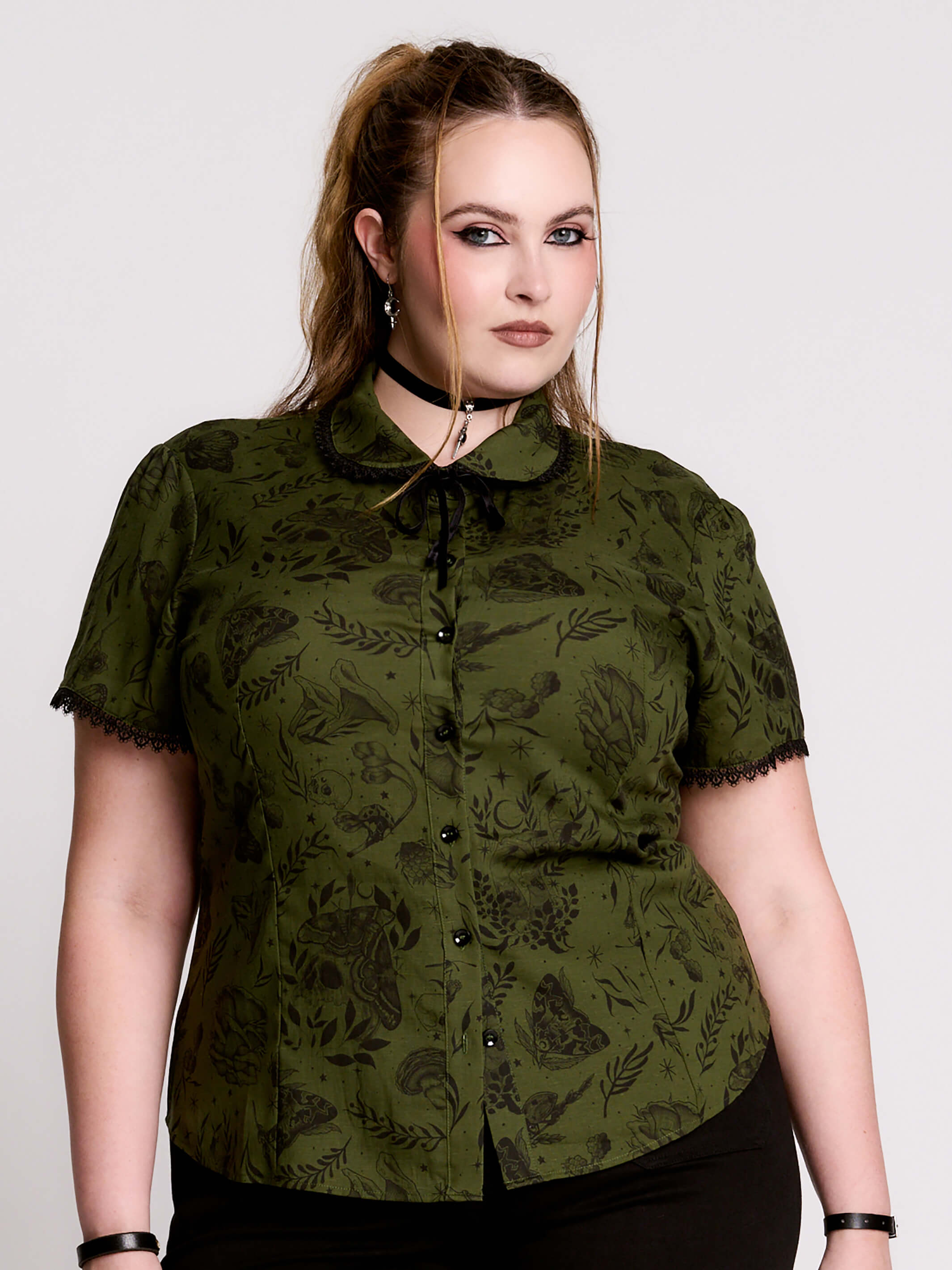 a short-sleeve olive green blouse with a dark botanical pattern, black lace trim collar with black ribbon tiedetail and black buttons. 